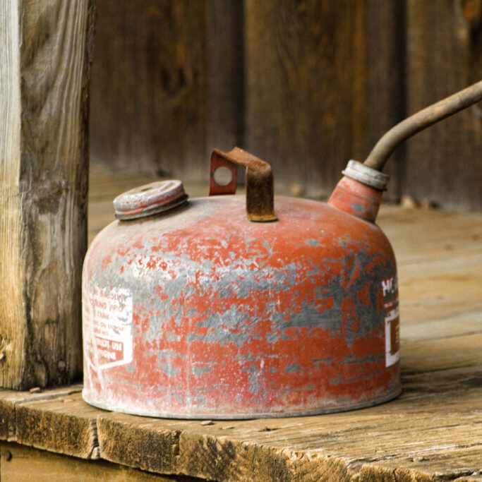 This is a old grungy painted red metal gas can sitting on a wood porch.